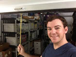 Kyle Messemore smiling as he pulls closed the door of the LGBTQ History Museum's storage unit. The unit behind him is full of shelves and archival boxes.